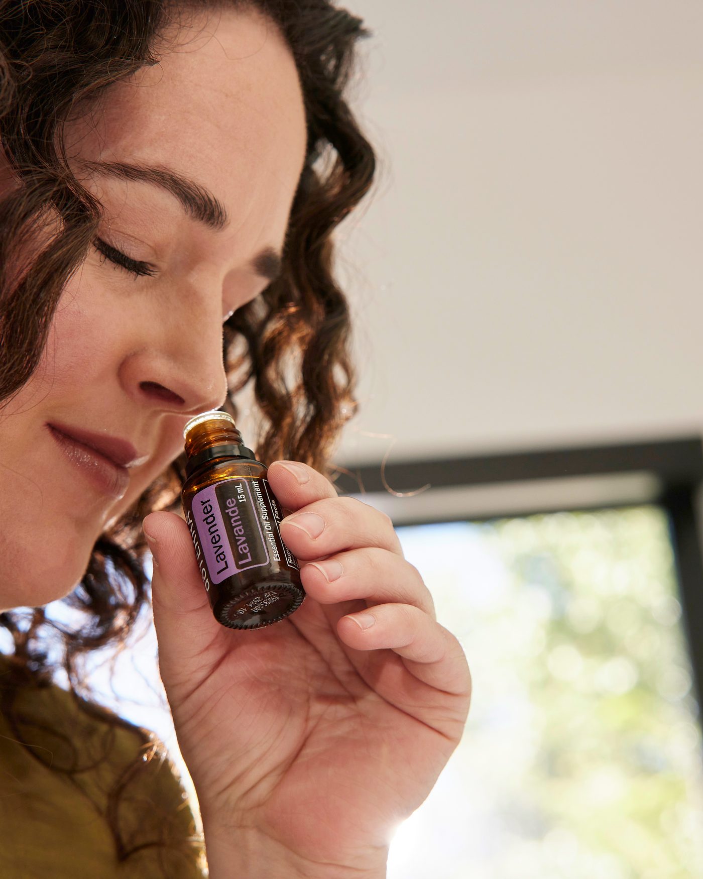 A woman smelling a bottle of dōTERRA Lavender essential oil by a sunny window