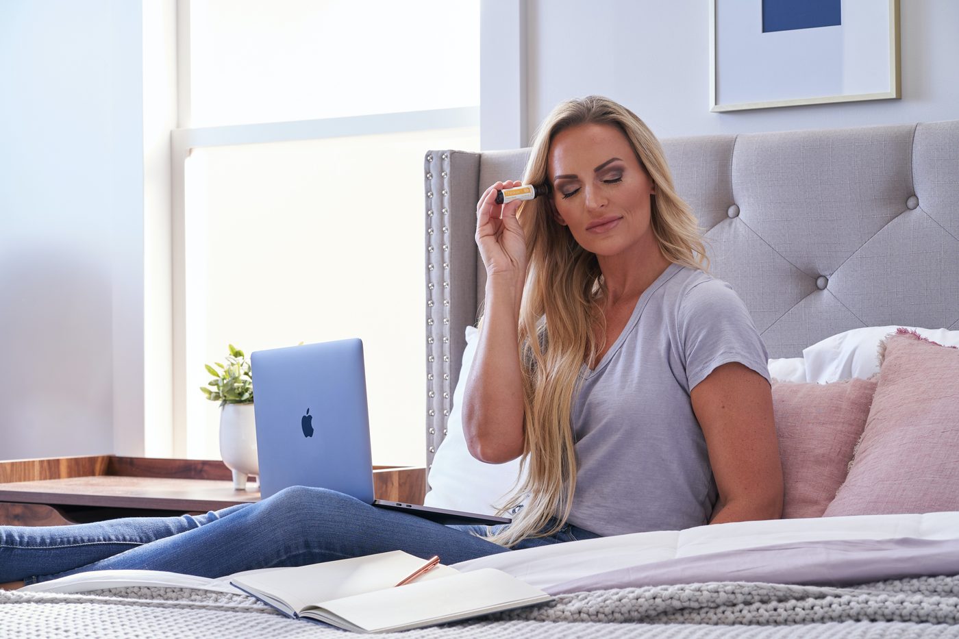 Une femme assise sur un lit avec un ordinateur portable, qui sent un flacon d'huile essentielle